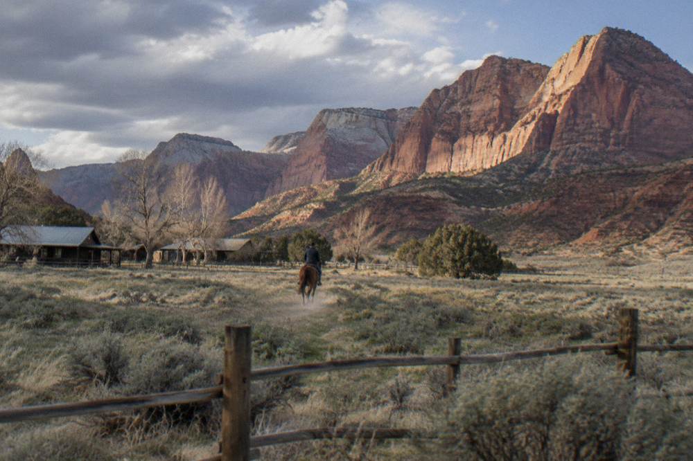 A rider on horseback crossing open sagebrush plateau toward canyon cliffs