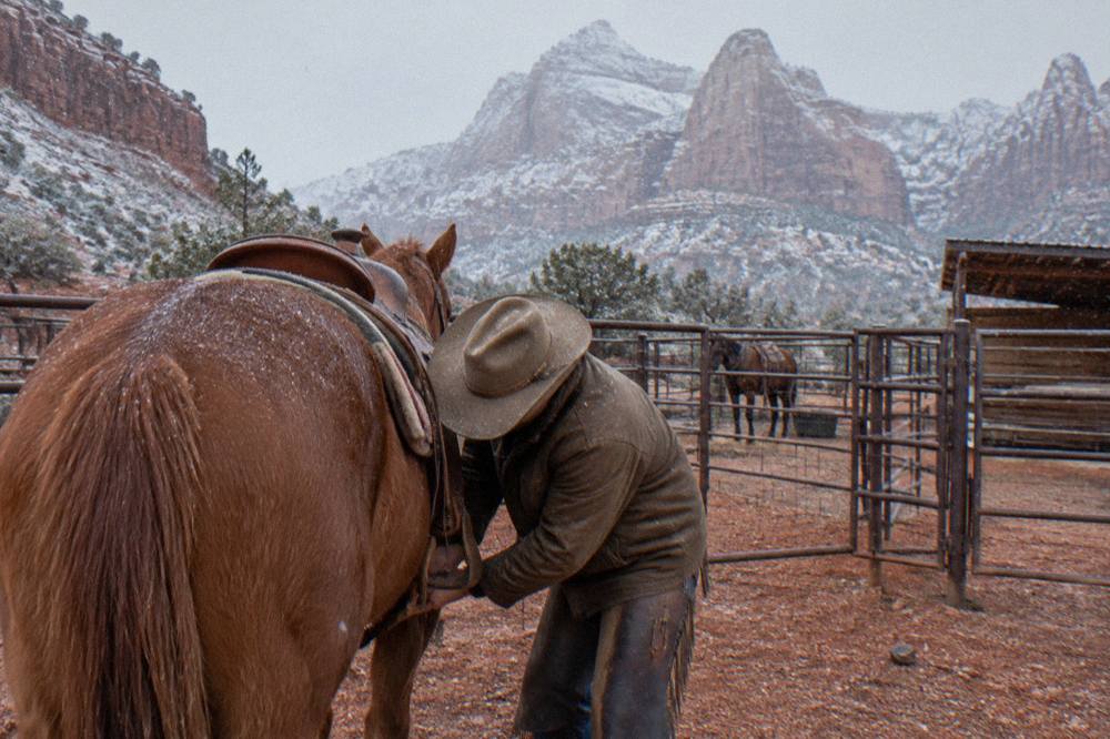 A wrangler adjusting a saddle cinch on a horse at a ranch before a ride
