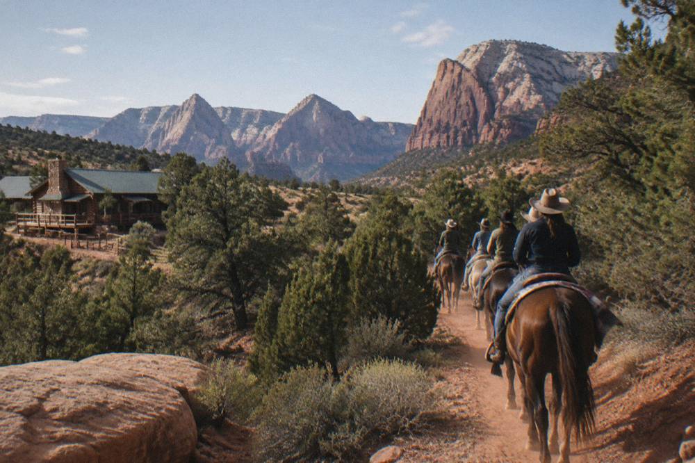 Horses and riders walking a red dirt trail between juniper trees and sandstone ledges