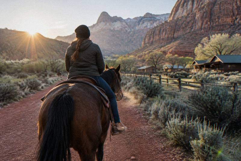 Riders on horseback crossing a golden meadow at sunrise beneath sandstone cliffs in southern Utah