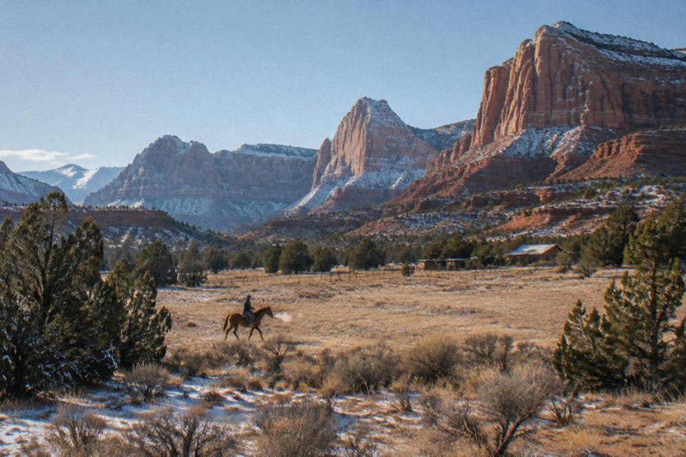 Lone rider on a horse crossing open high desert ground toward red mesas