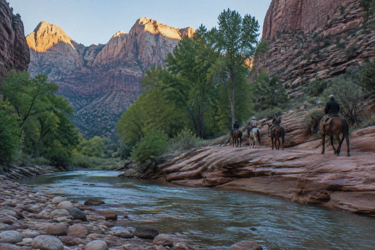 Horseback riders on a sandstone trail beside a river in a narrow desert canyon