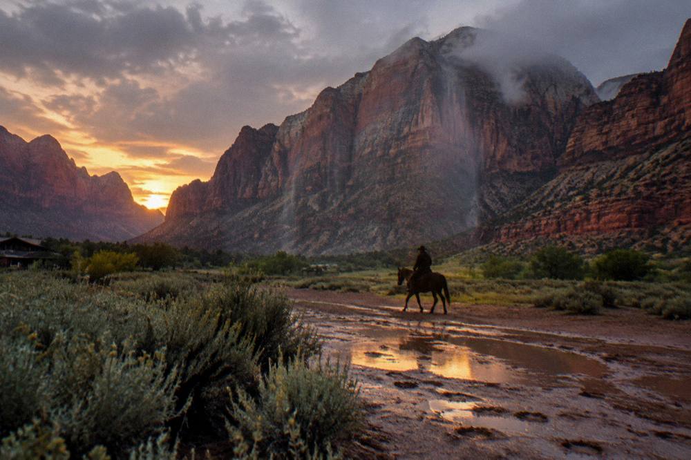 Rider on horseback silhouetted against wide canyon walls at sunset