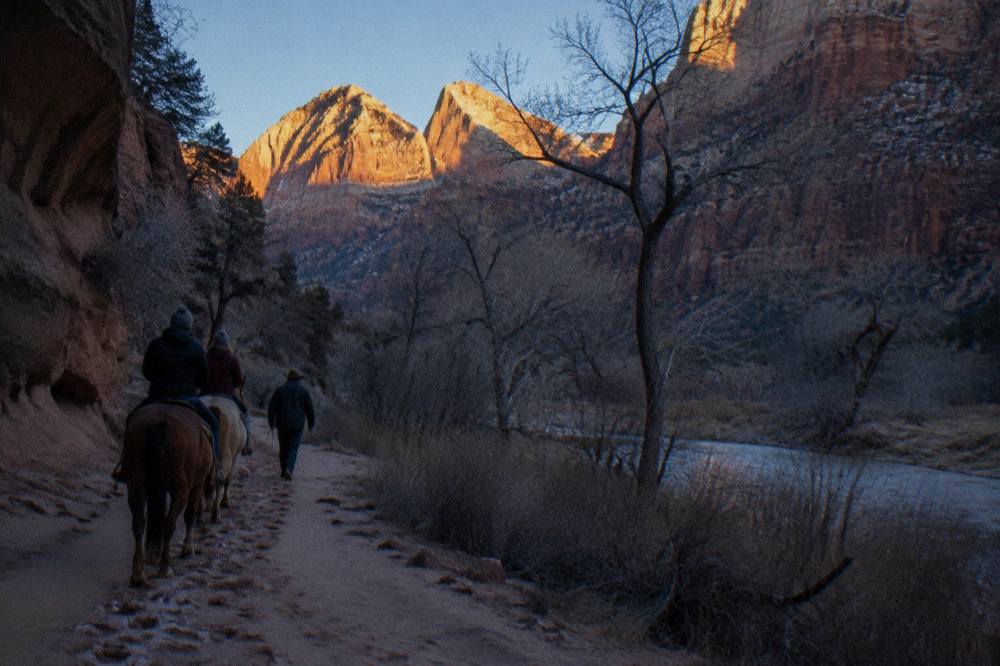 A guide leading two riders on horseback along a sandy trail between tall red rock canyon walls.