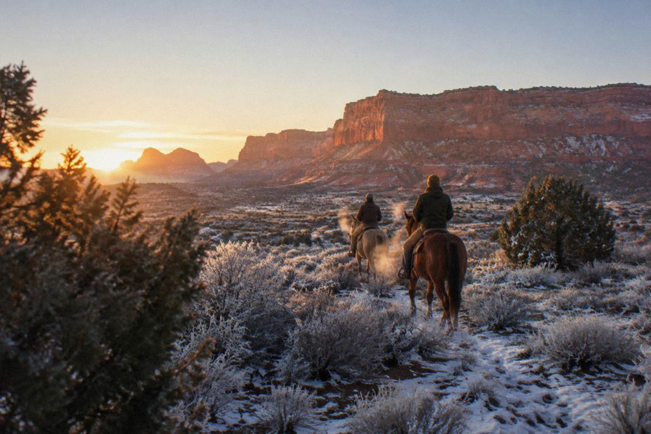 Two riders on horseback crossing open high desert rangeland with red rock mesas in the distance at golden hour.