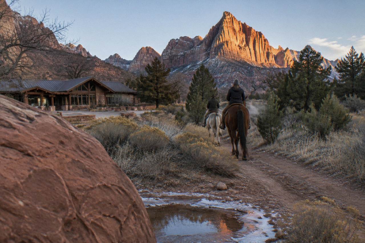 Two riders on horseback following a dirt trail through open high-desert country near Zion