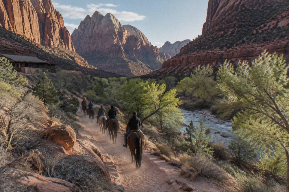 A string of riding horses on a sandy bench trail with layered red rock walls rising above