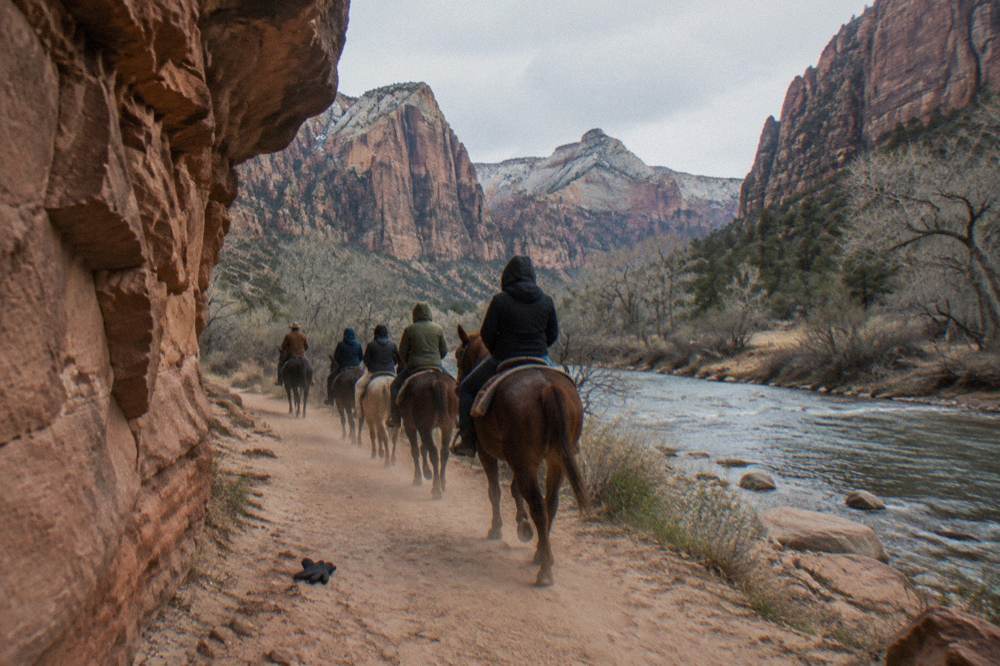 A wrangler leading a line of riders along a flat trail between river and canyon wall