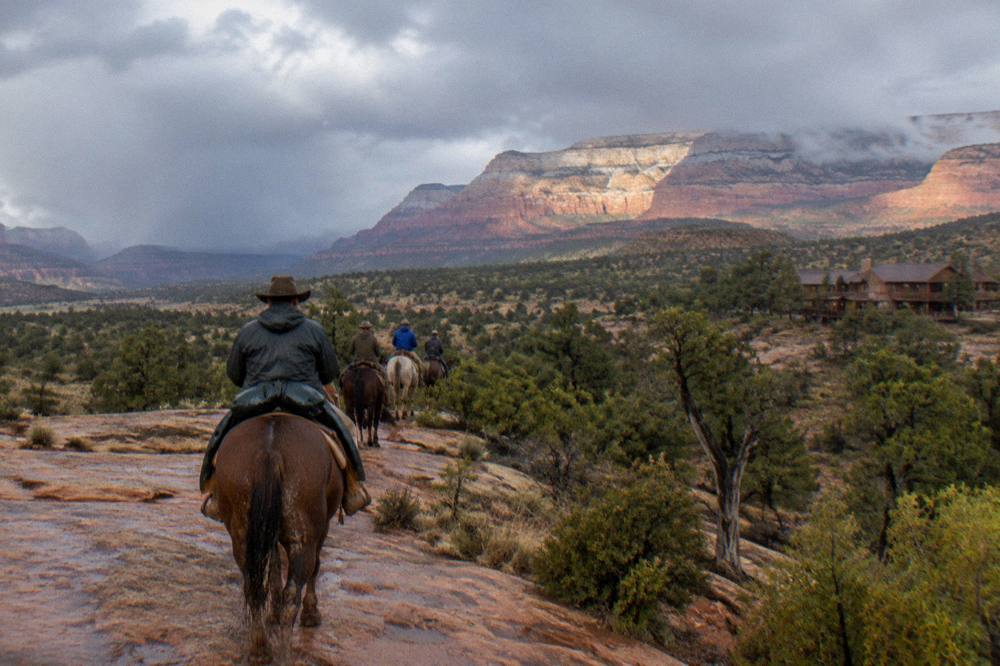 Riders on horseback crossing open juniper-dotted high country with layered cliffs in the distance