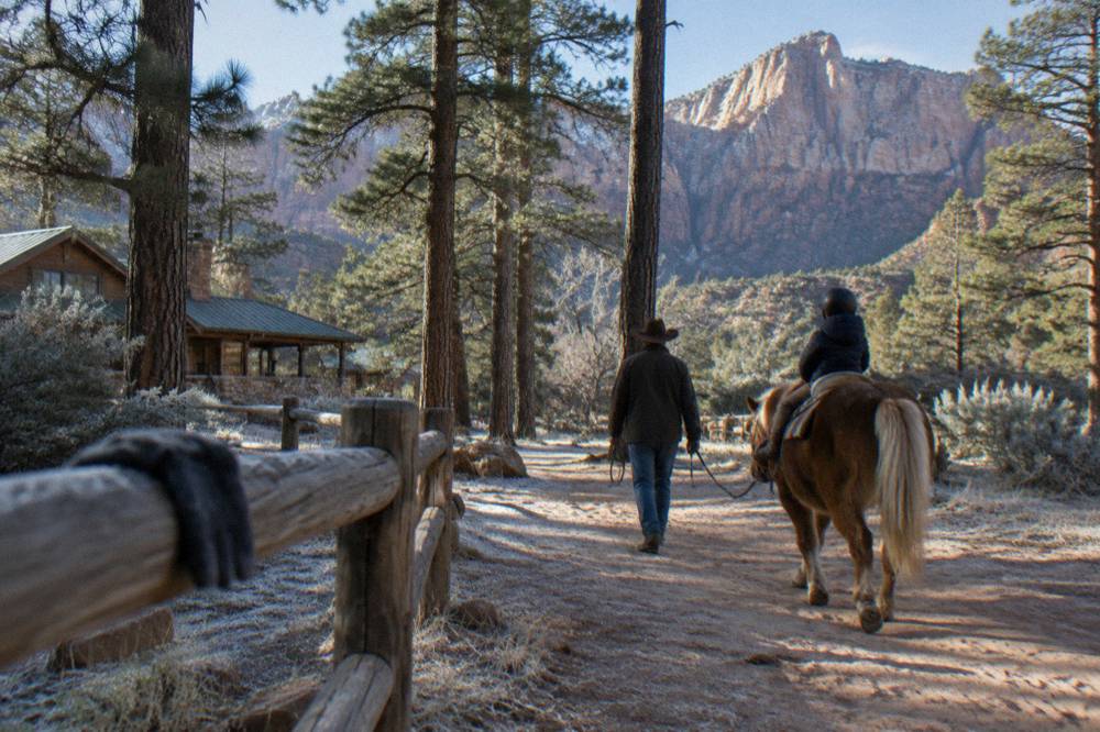 A wrangler on foot leading a saddled horse with a small child rider along a sandy path