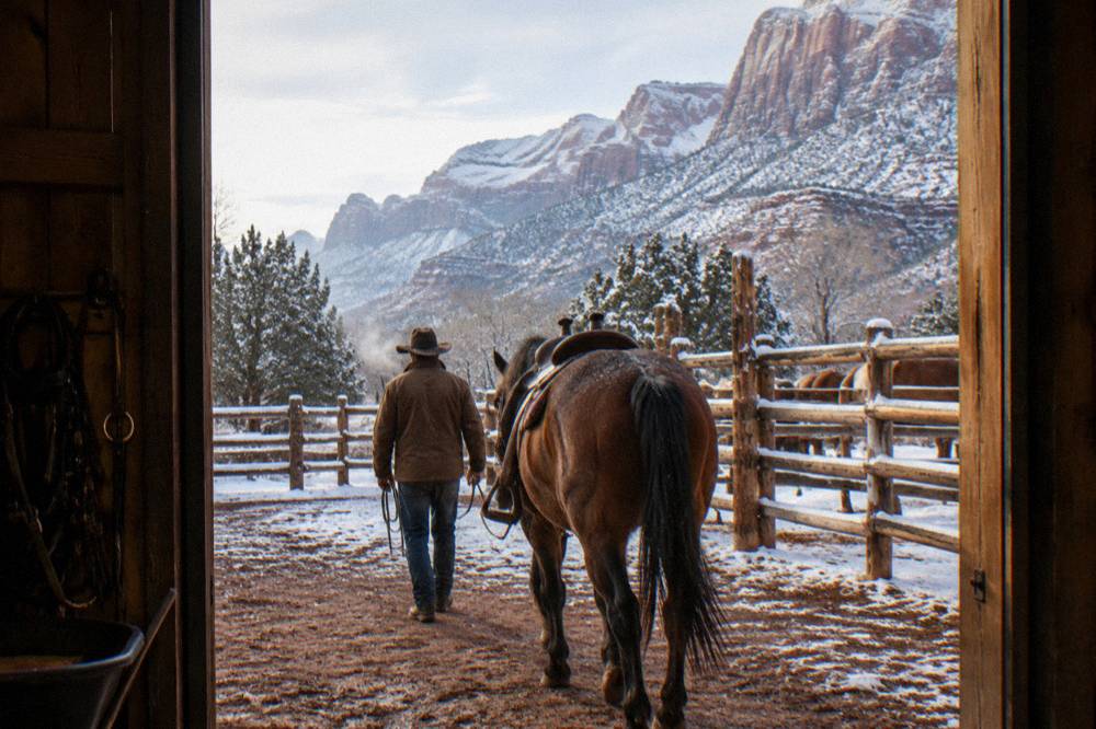 Wrangler in a cowboy hat leading a saddled horse by the reins near wooden corrals