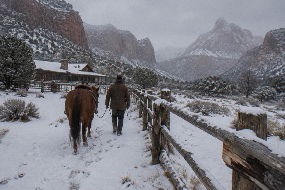 A wrangler leading a quiet horse by the reins along a fenced ranch path
