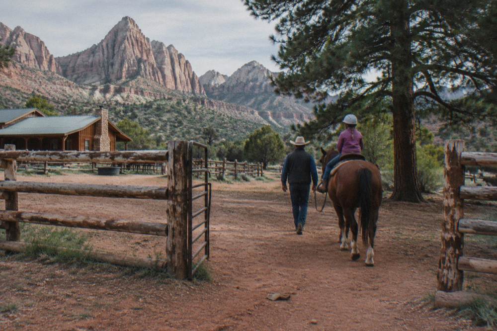A wrangler leading a child on a quiet horse along a ranch fence line