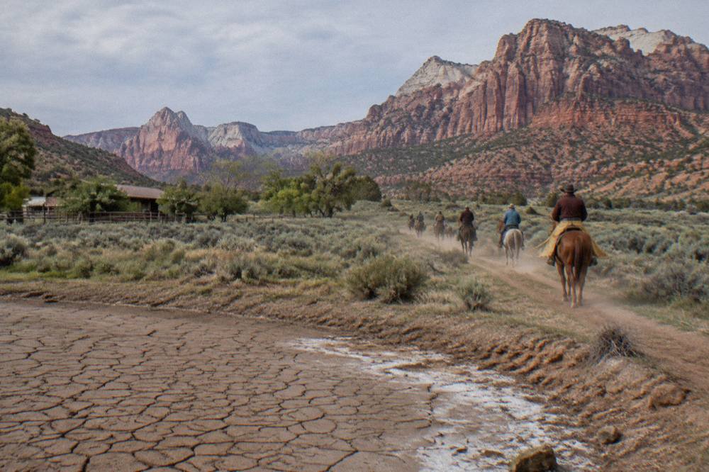 Horses and riders on a dirt trail crossing open sage country toward red rock cliffs
