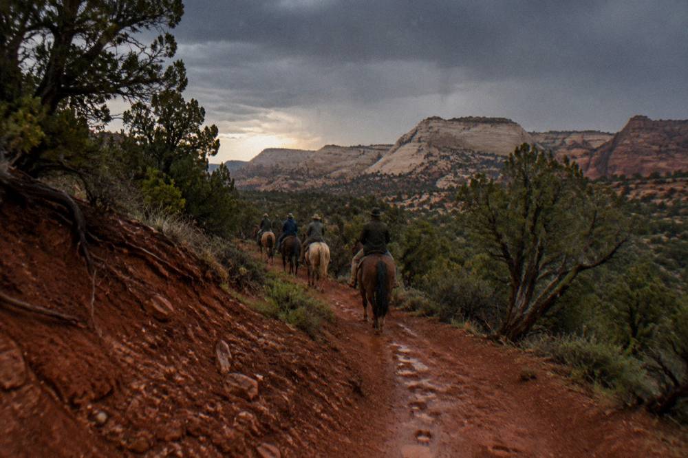 Riders mounted in a line crossing a red dirt track between juniper trees and pale rock hills