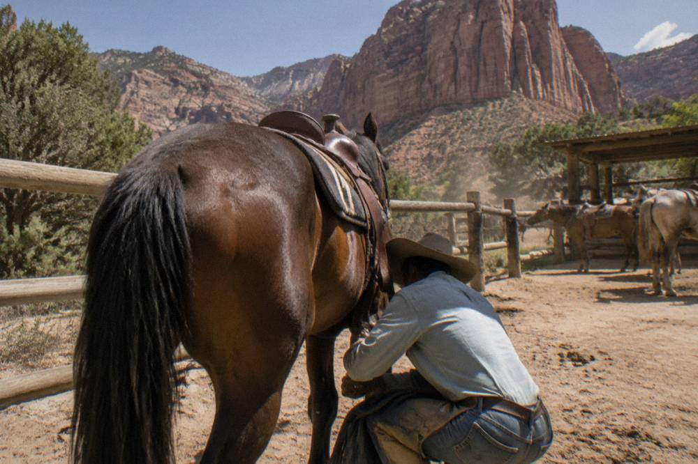 A wrangler checking the girth strap on a saddled horse beside a red rock butte