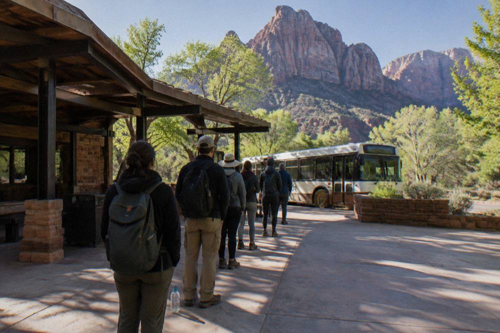 A line of visitors waiting at an outdoor shuttle stop beside red rock walls in morning light.