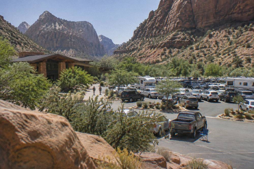 A filled parking lot with cars lined up near a visitor center building at the base of cliffs.