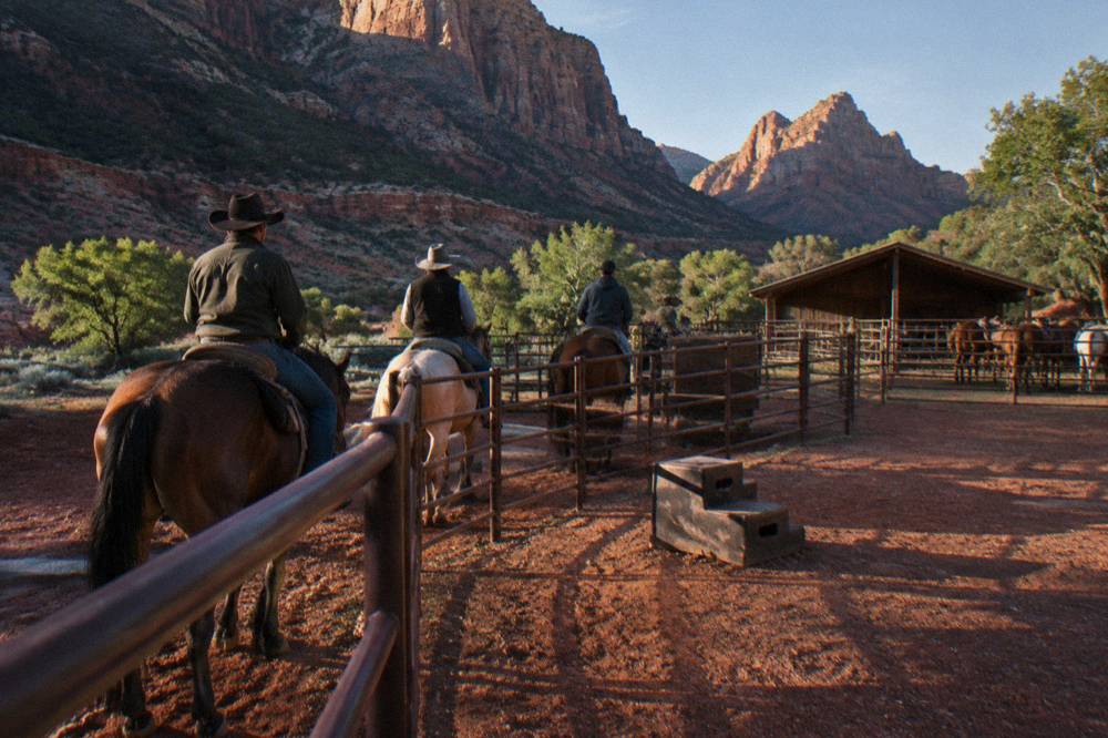 Riders mounting horses at a corral fence in late afternoon shade