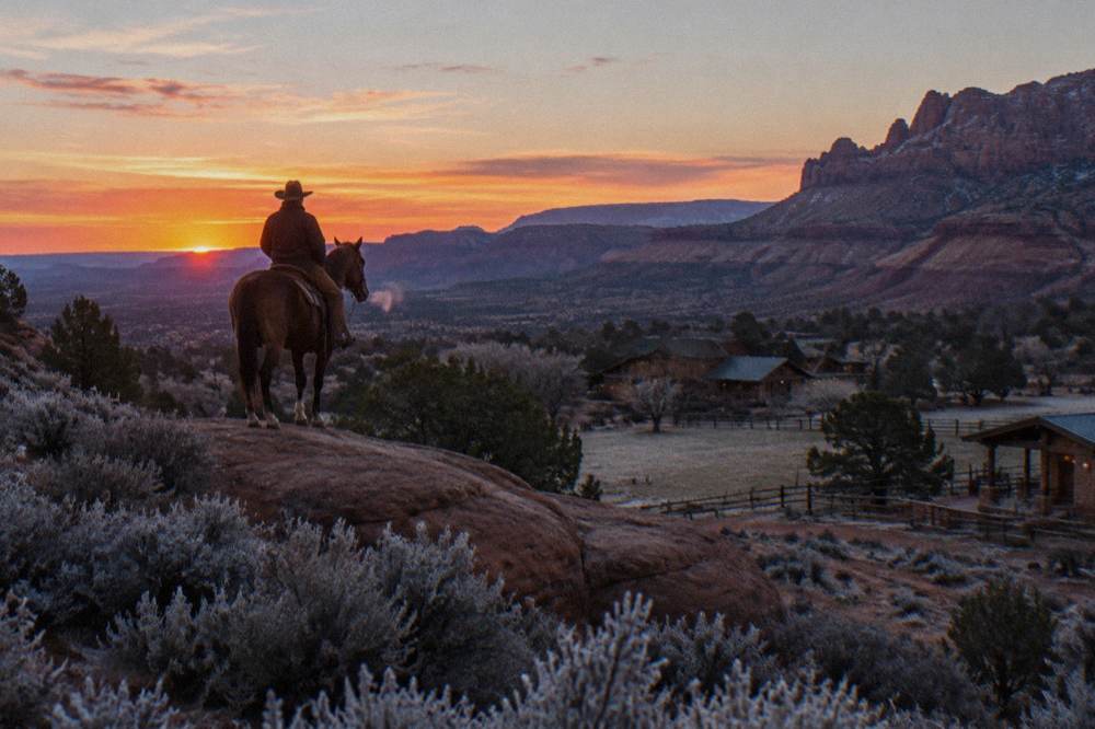 A rider on horseback silhouetted against a wide sunrise sky over layered mesas